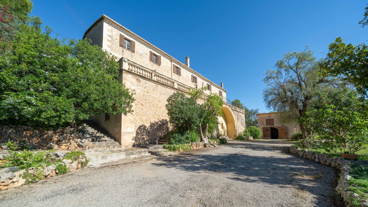 Grande villa en pierre avec terrasse surélevée, volets en bois et arbres sous un ciel bleu clair.