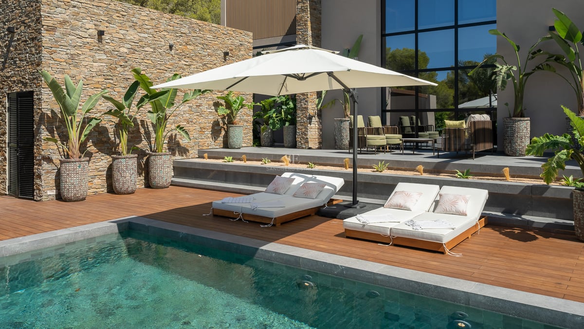 Piscine moderne avec terrasse en bois, bains de soleil blancs et parasol devant un mur en pierre naturelle.