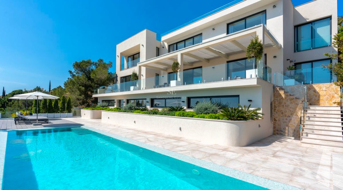 Grande piscine turquoise devant une villa blanche moderne de trois étages avec balcons en verre et escaliers en pierre.