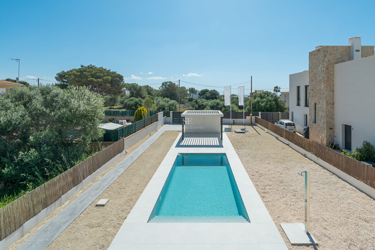 Une longue piscine rectangulaire aux eaux turquoise, entourée de dalles blanches, de gravier et d'une villa contemporaine.