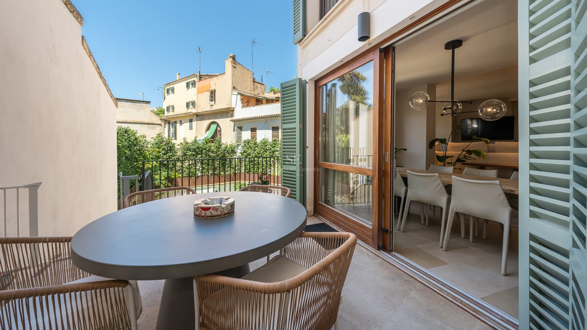 Terrasse avec table ronde grise, chaises en corde tressée et baies vitrées ouvrant sur le séjour.