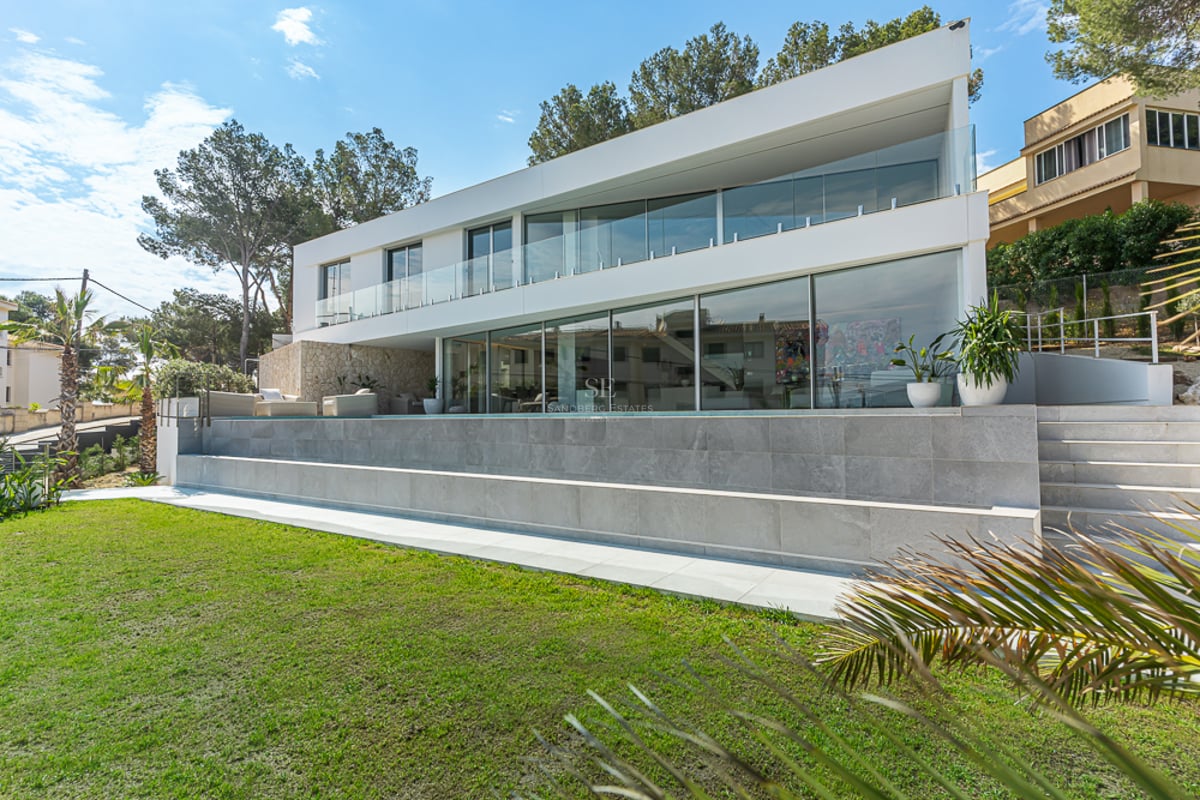 Villa moderne blanche avec grandes baies vitrées, piscine à débordement et pelouse verte sous un ciel bleu.