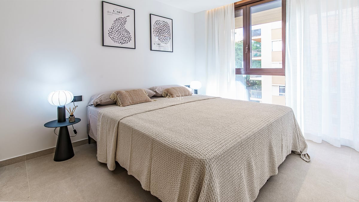 Chambre moderne avec grand lit, linge de lit beige, lampes minimalistes et grandes fenêtres avec voilages blancs.