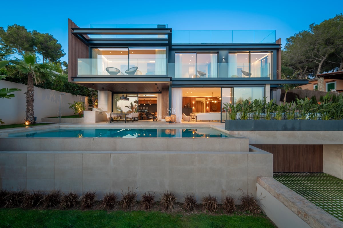 Villa moderne de deux étages avec balcons en verre et piscine à débordement éclairée au crépuscule.