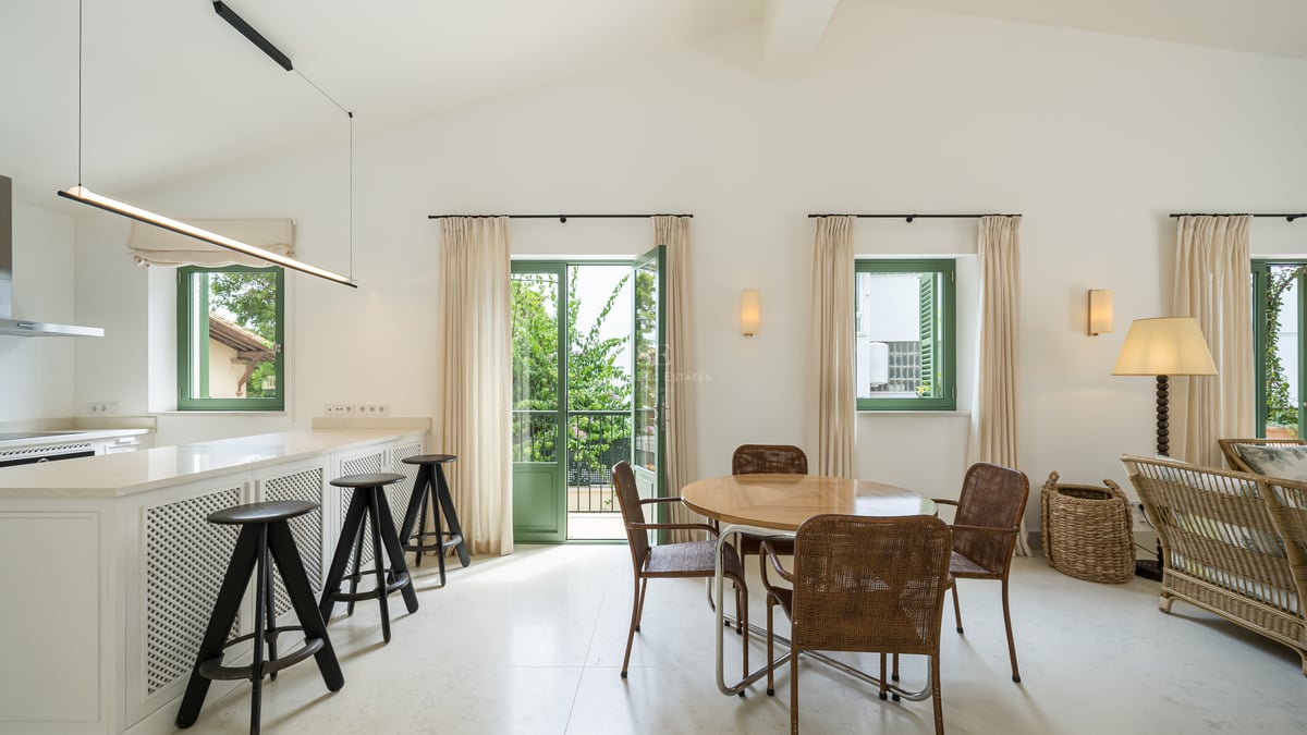 Salle à manger moderne avec table ronde en bois, chaises tressées et portes-fenêtres vertes ouvrant sur un jardin.