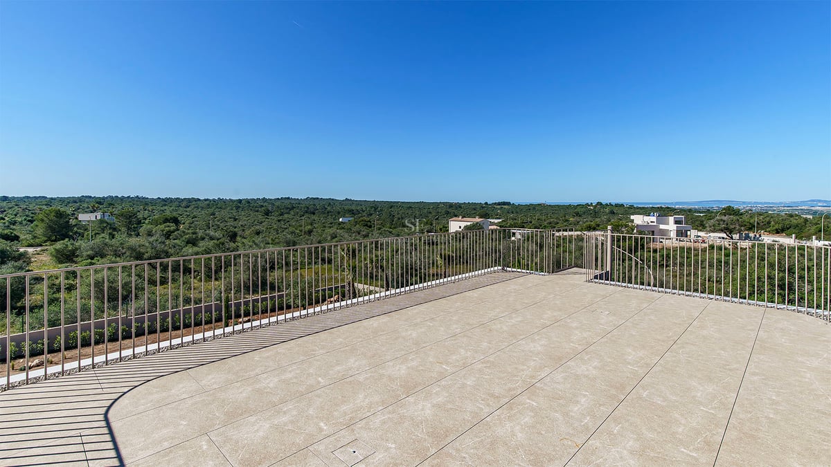 Terrasse en pierre avec garde-corps métallique surplombant un paysage verdoyant sous un ciel bleu azur.