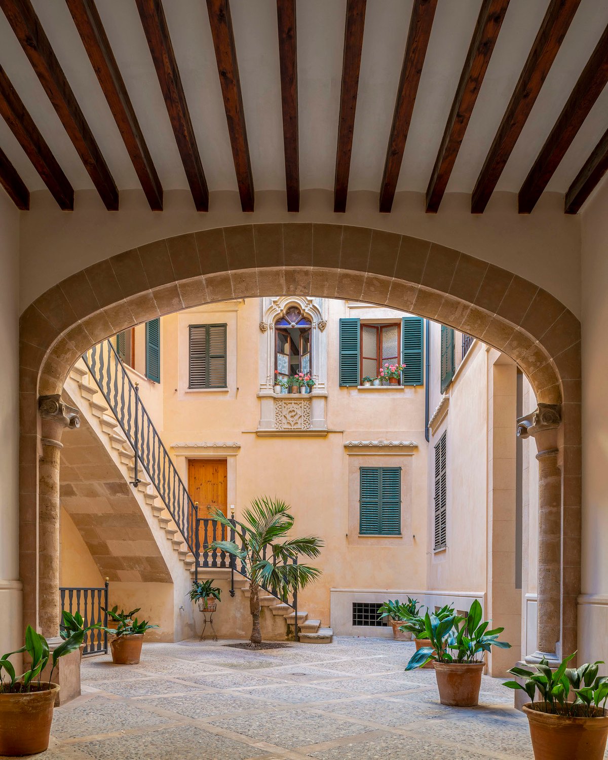Cour intérieure avec arche en pierre, poutres en bois, escalier en pierre et plantes en pots sous lumière naturelle.