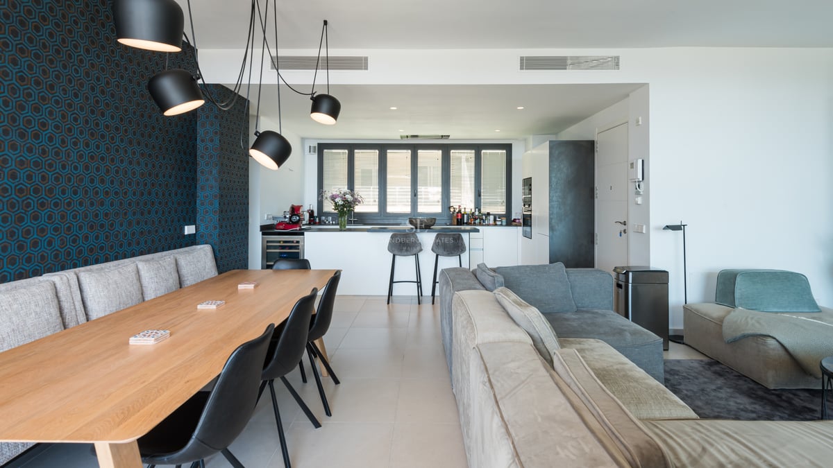 Pièce à vivre ouverte comprenant une table en bois, des canapés gris, un mur géométrique bleu et une cuisine moderne.