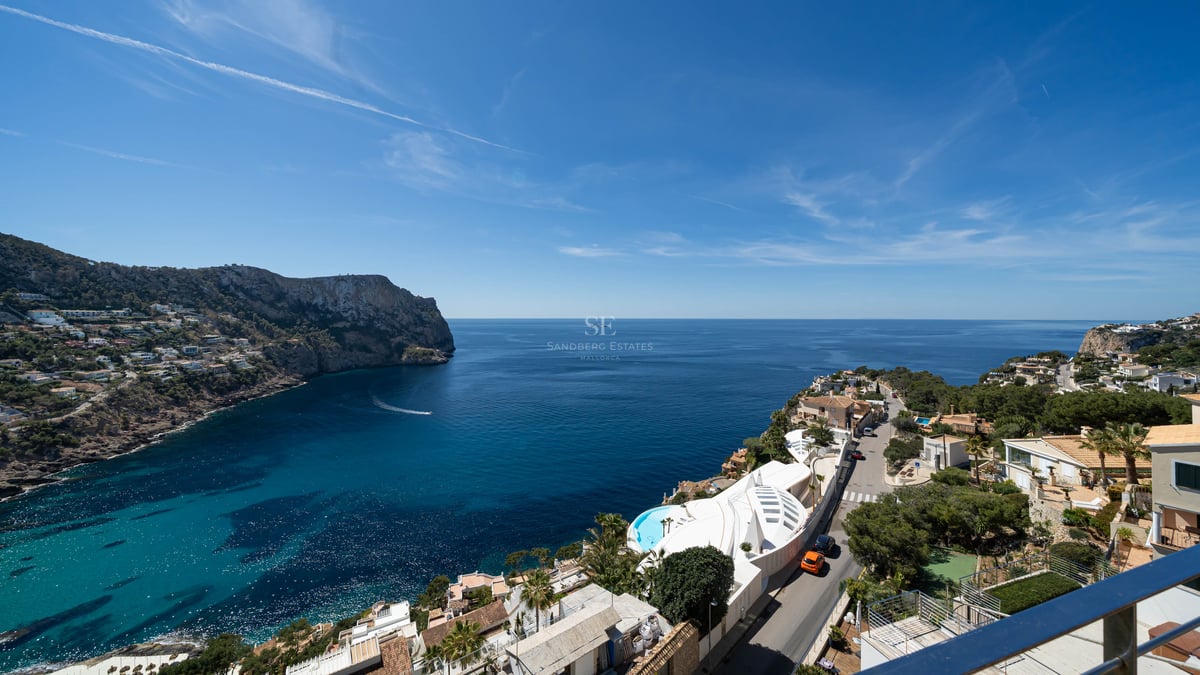 Vue en hauteur d'une baie bleu azur entourée de falaises rocheuses et de villas de luxe sous un ciel dégagé.