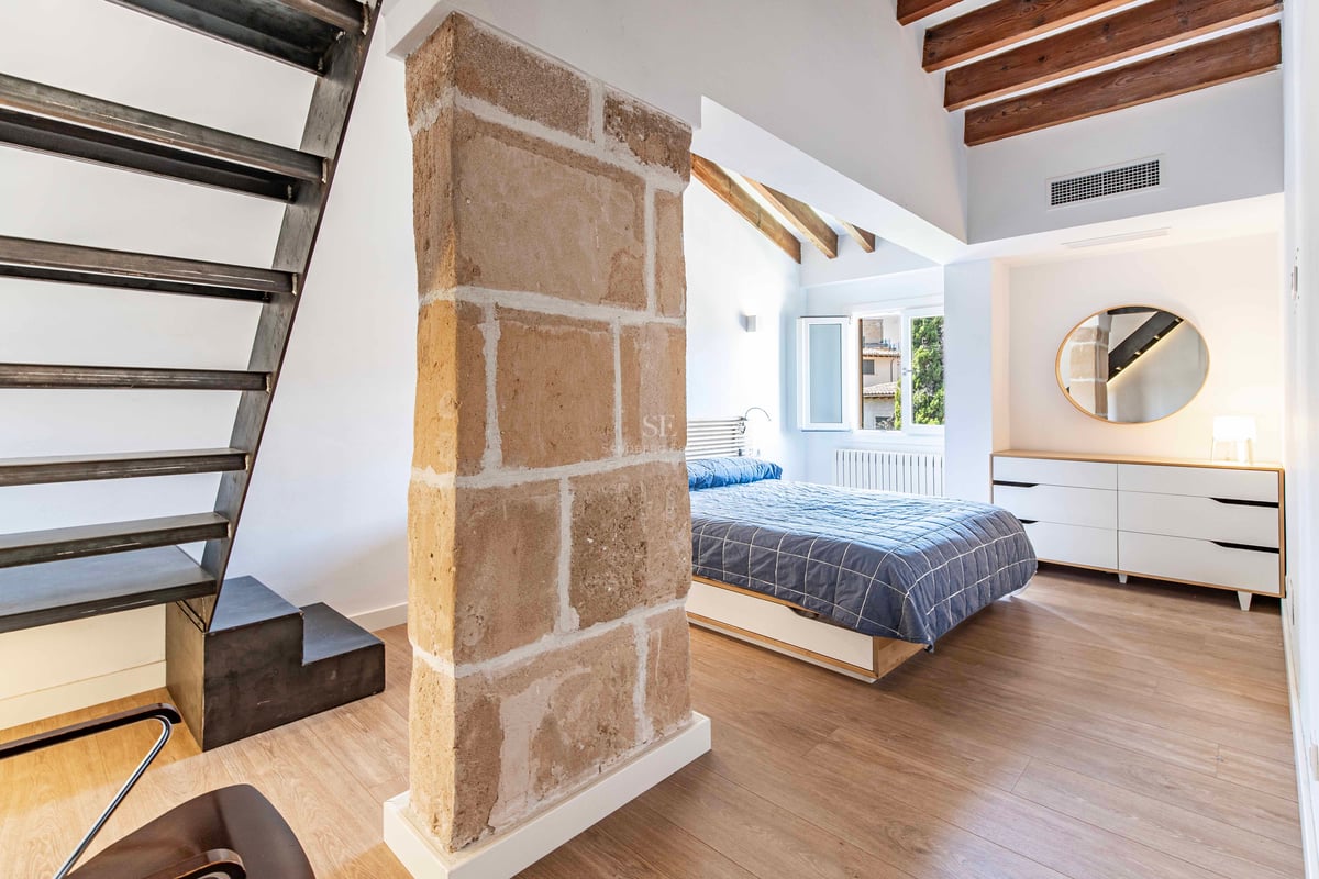 Chambre lumineuse avec pilier en pierre naturelle, escalier en acier industriel, poutres en bois et lit double.