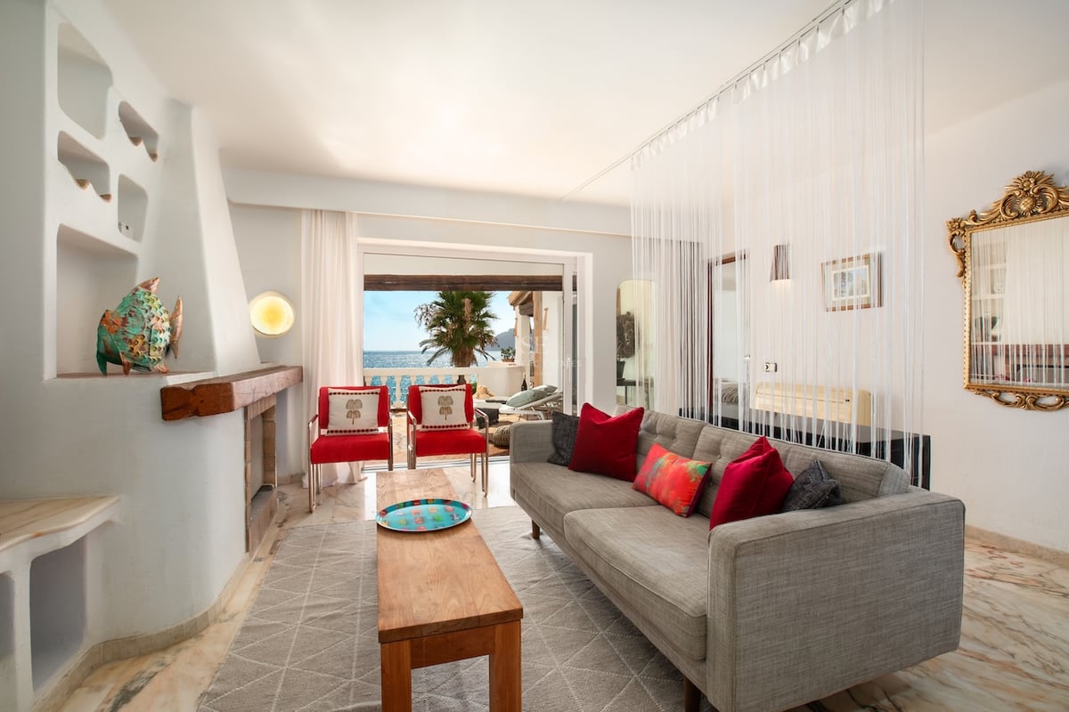 Salon avec canapé gris, chaises rouges, cheminée rustique et grandes baies vitrées ouvrant sur la mer et une terrasse.