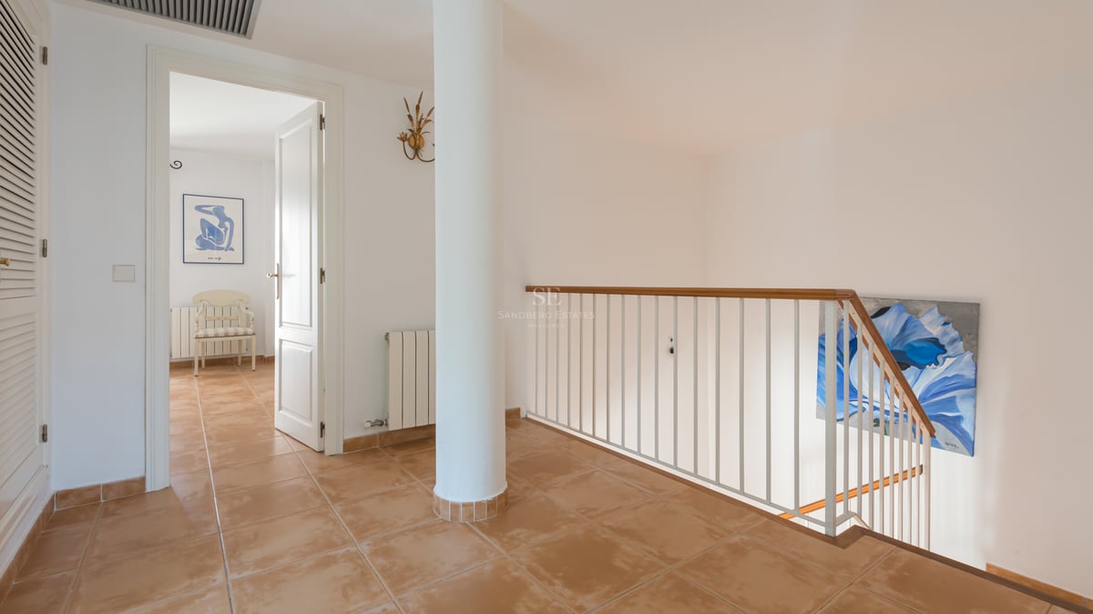 Couloir avec carrelage en terre cuite, pilier blanc et rampe d'escalier menant à une porte ouverte.