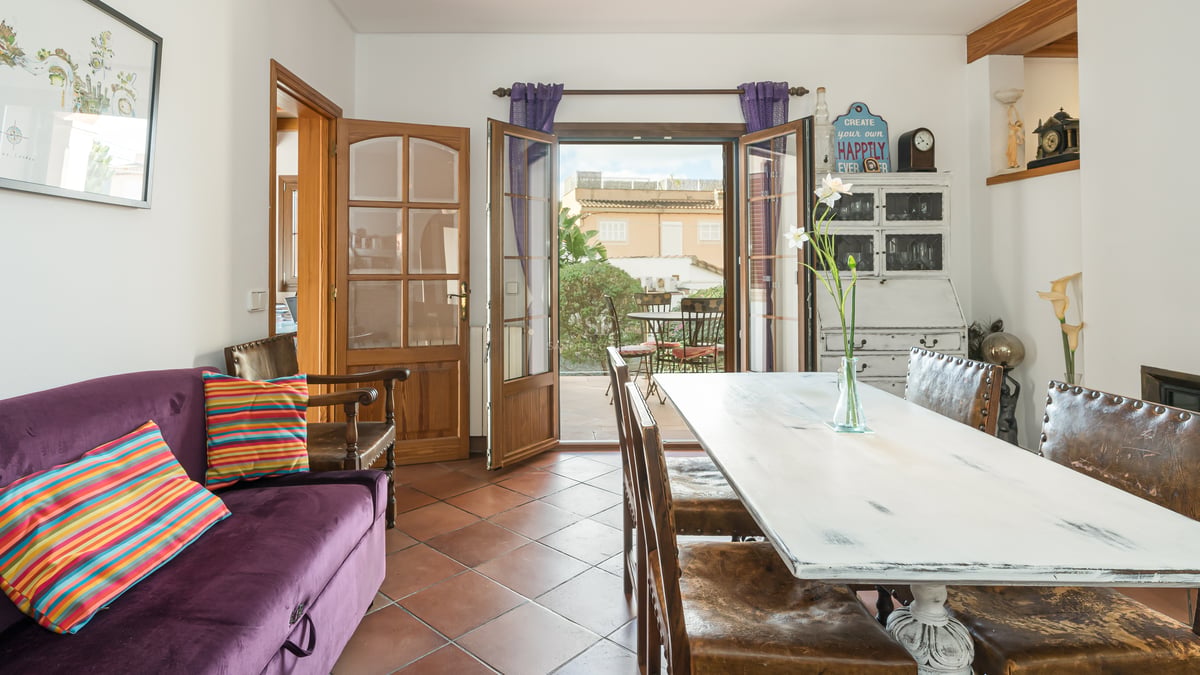 Salle à manger avec sol en terre cuite, table en bois blanc vieilli et portes ouvertes sur un patio ensoleillé.