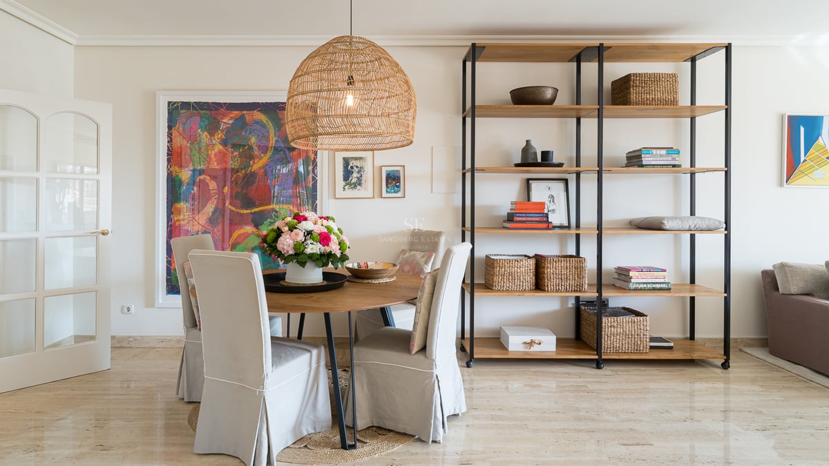 Salle à manger avec table ronde en bois, chaises beiges, suspension en rotin et étagère moderne sur sol en marbre.