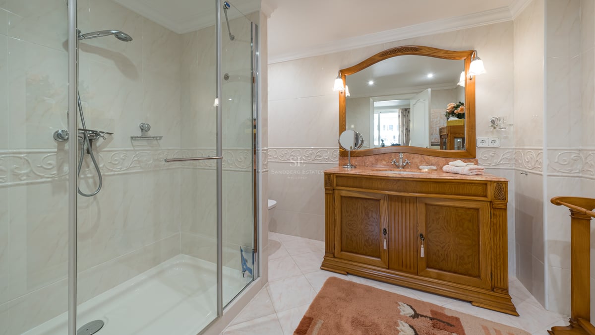 Salle de bain classique avec douche en verre, carrelage en marbre crème et un grand meuble-lavabo en bois sculpté.
