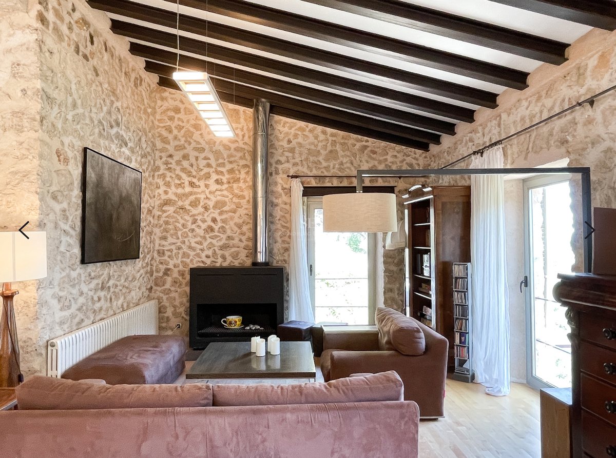 Salon avec murs en pierre, plafond à poutres sombres, cheminée moderne et grandes fenêtres avec lumière naturelle.