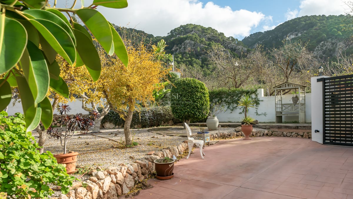 Terrasse en terre cuite et jardin avec murs en pierre et arbres matures sur fond de montagnes rocheuses.