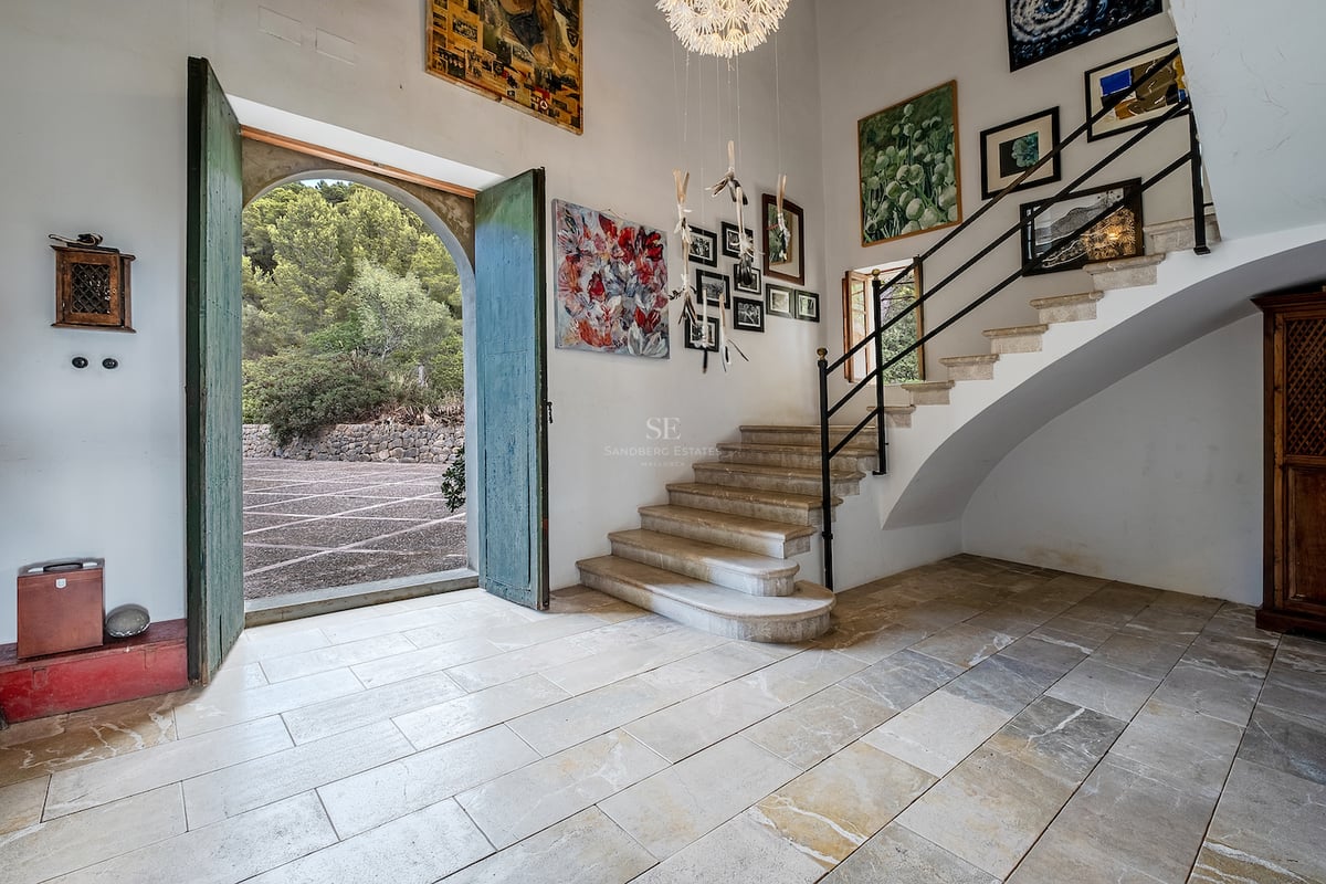 Vaste hall d'entrée avec sols en pierre, hauts plafonds, escalier en pierre et grandes portes vertes ouvertes.