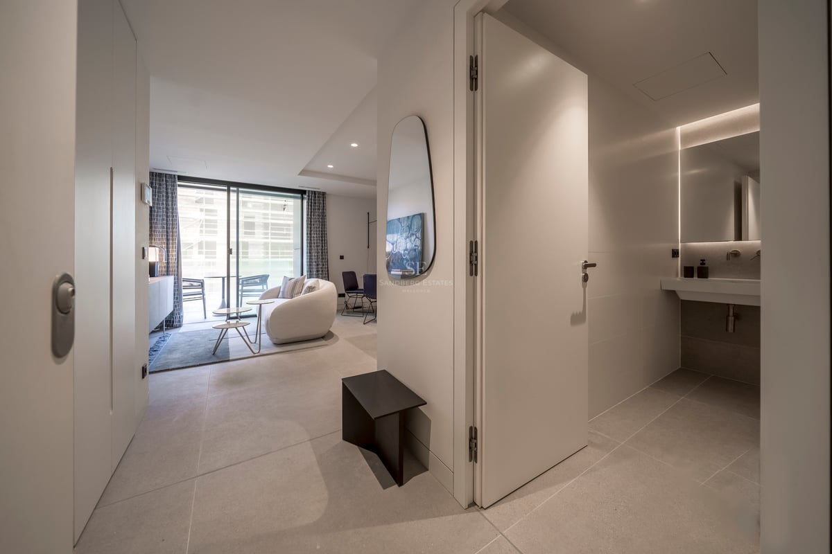 Couloir moderne menant à un salon lumineux avec canapé incurvé et une salle de bain chic sur la droite.