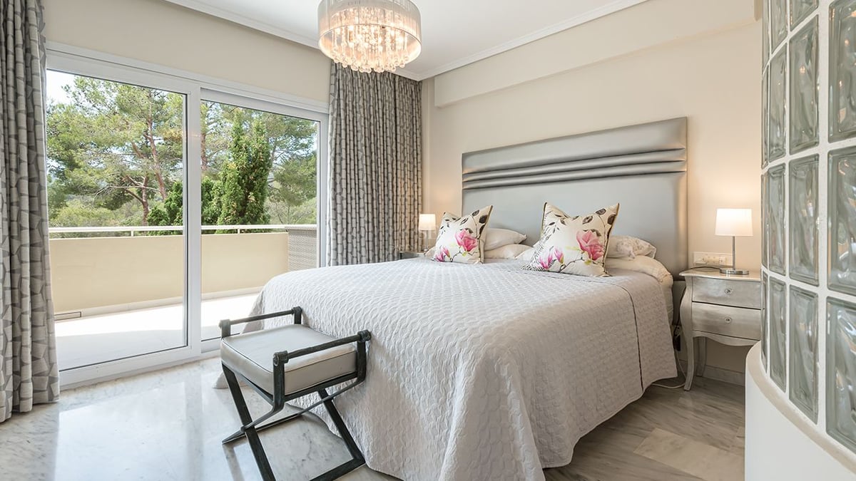 Chambre principale lumineuse avec sol en marbre, tête de lit argentée et accès terrasse vue sur les arbres.