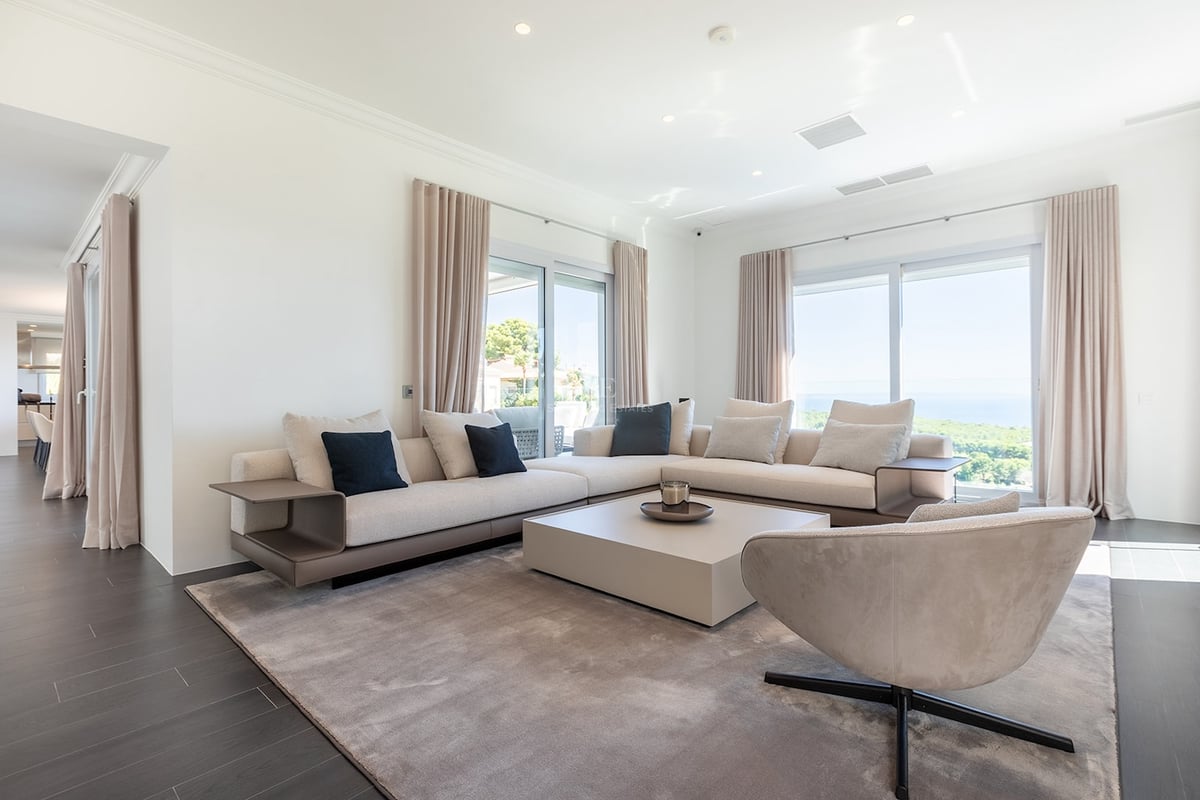 Salon blanc spacieux avec canapé d'angle beige, fauteuil design et grandes baies vitrées donnant sur l'océan.