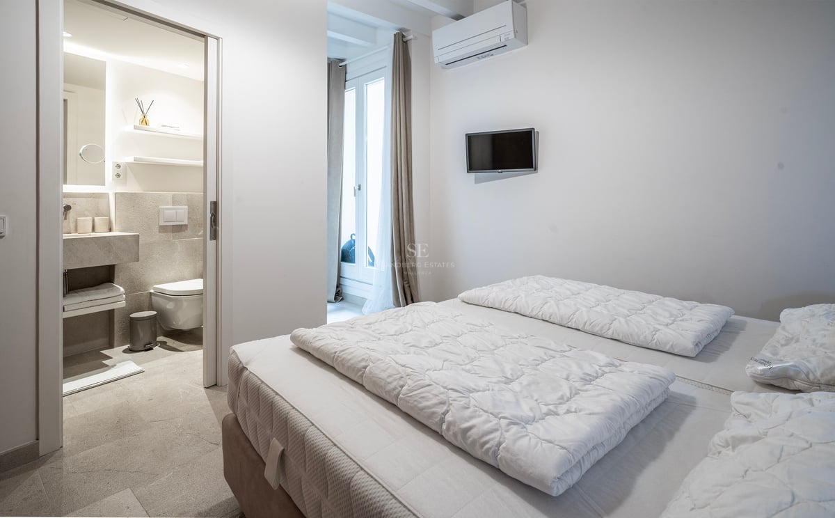 Chambre lumineuse avec literie blanche et porte ouverte sur une salle de bain moderne en pierre.
