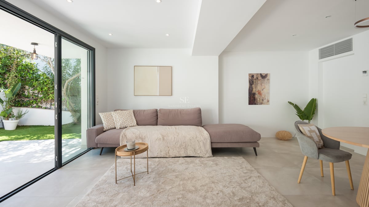 Salon moderne avec canapé mauve, tapis moelleux et baies vitrées s'ouvrant sur un jardin verdoyant.