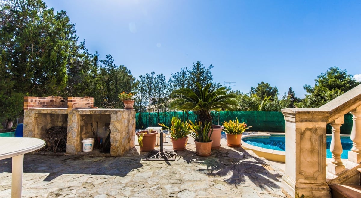 Terrasse en pierre ensoleillée avec piscine bleue, barbecue intégré et plantes en pots sous un ciel dégagé.