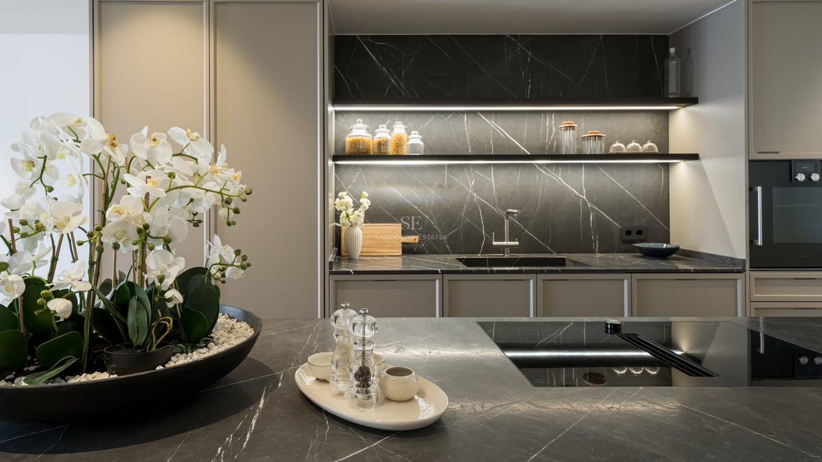 Modern kitchen featuring dark marble countertops, integrated appliances, and warm LED shelf lighting with white orchids.