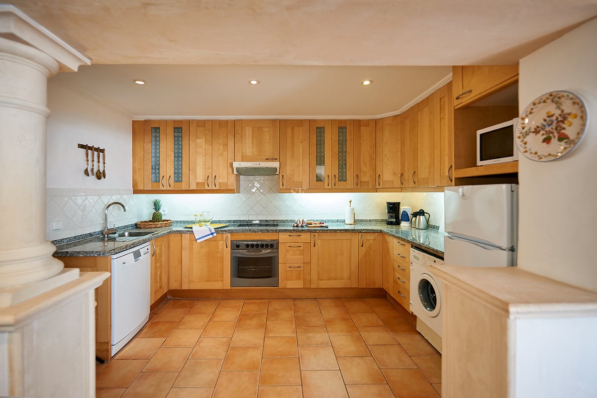 Cuisine rustique avec armoires en bois clair, plans de travail en granit, sol en terre cuite et électroménager.