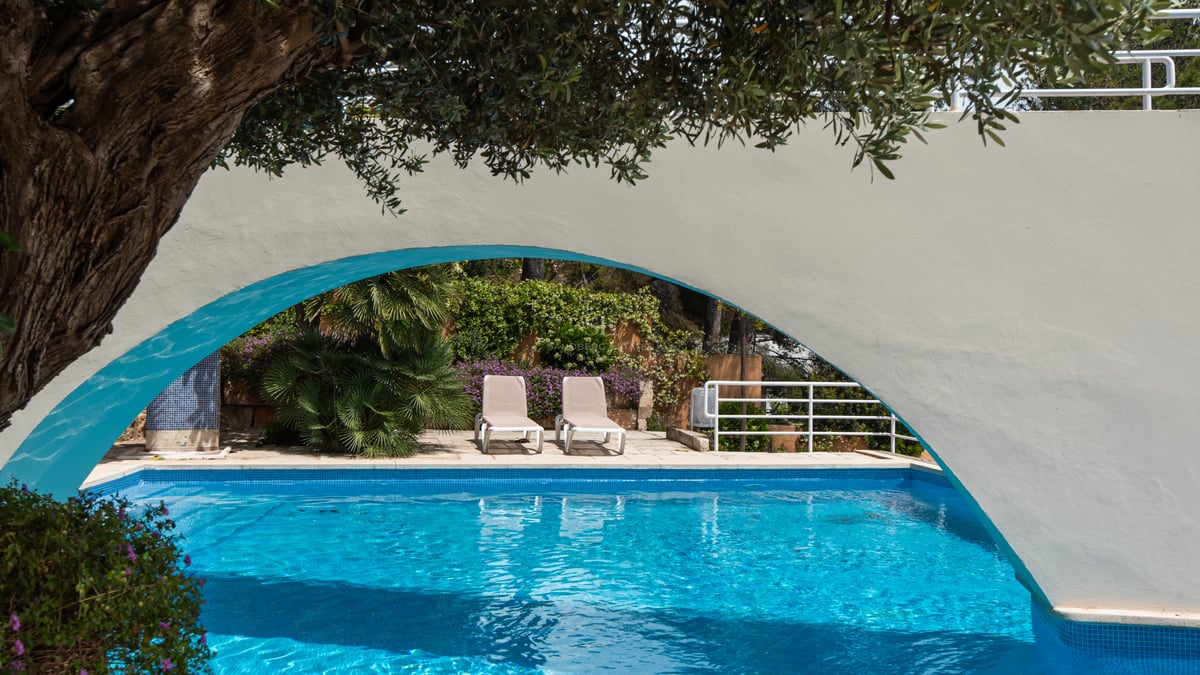 Piscine bleue et transats encadrés par une arche blanche et une végétation luxuriante dans un jardin.