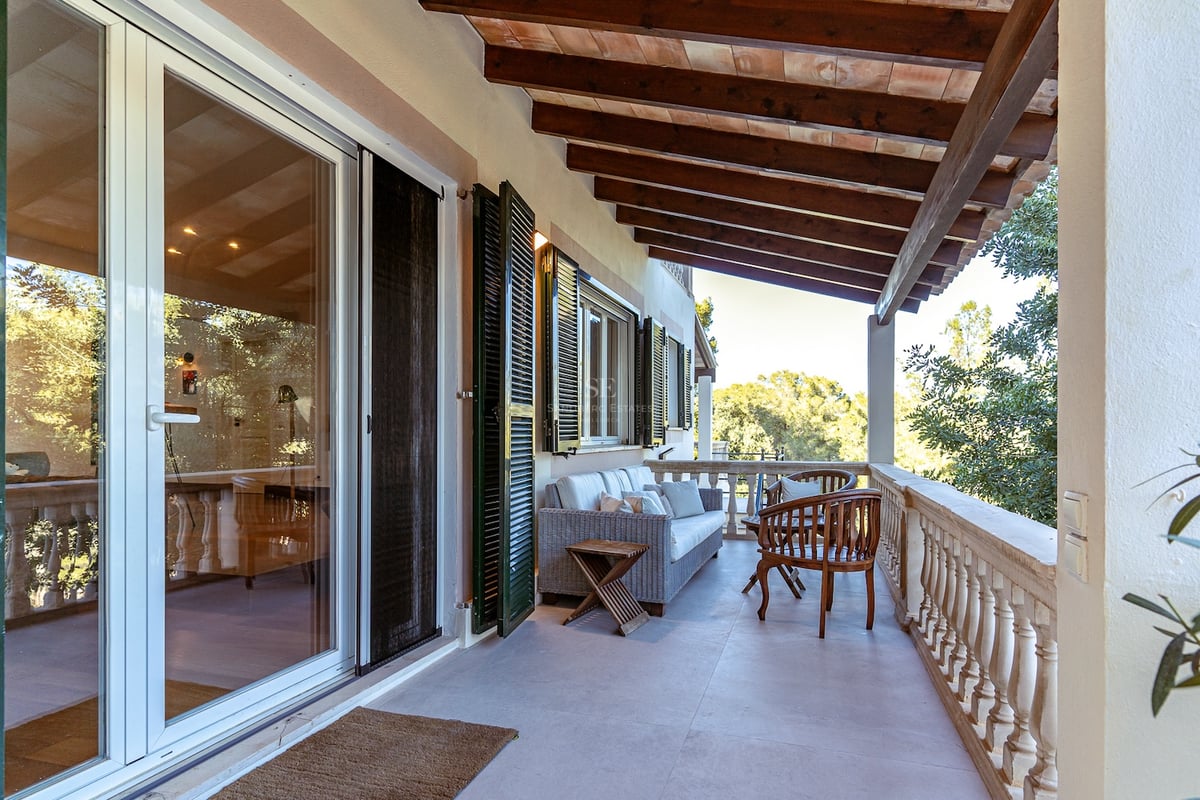 Terrasse couverte avec poutres apparentes, canapé en osier, balustrade en pierre et vue sur la verdure.