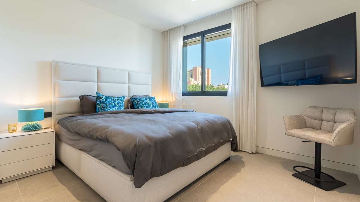 Chambre moderne avec grand lit, tête de lit rembourrée grise, TV murale et fenêtre lumineuse.