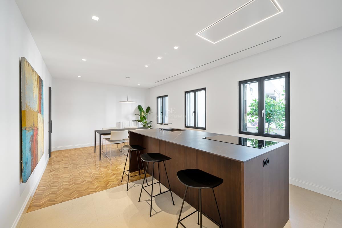 Cuisine minimaliste avec îlot central sombre, tabourets noirs, parquet à chevrons et tableau abstrait coloré.