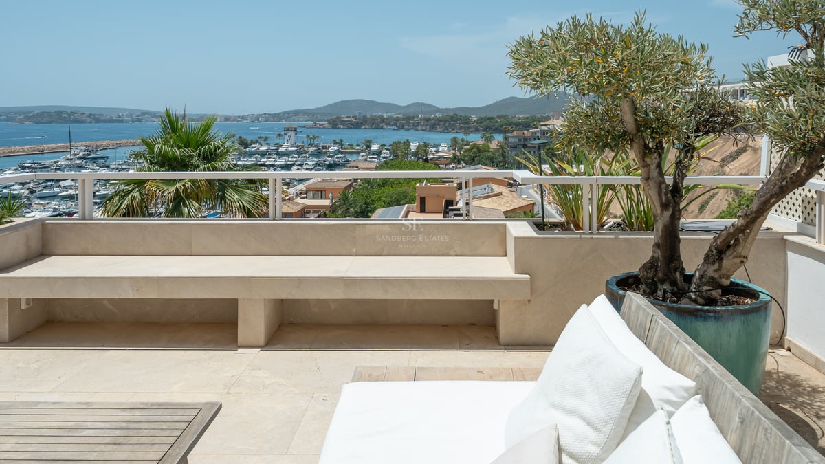 Canapé blanc et banc en pierre sur une terrasse surplombant un port rempli de yachts et des montagnes.