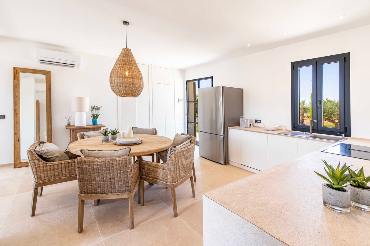 Bright dining area featuring a round wooden table, wicker chairs, a large woven pendant, and a modern kitchen with stone counters.