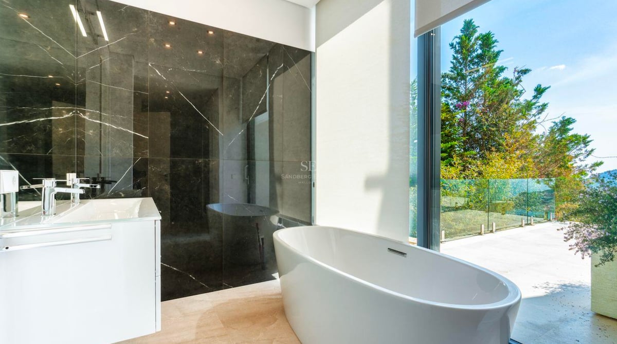 Salle de bain moderne dotée d'une baignoire îlot blanche devant une grande baie vitrée avec vue sur la verdure.