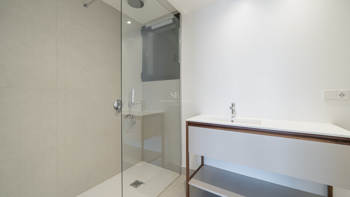 Baño moderno con ducha de cristal, azulejos beige y un mueble de lavabo elegante en blanco y madera.