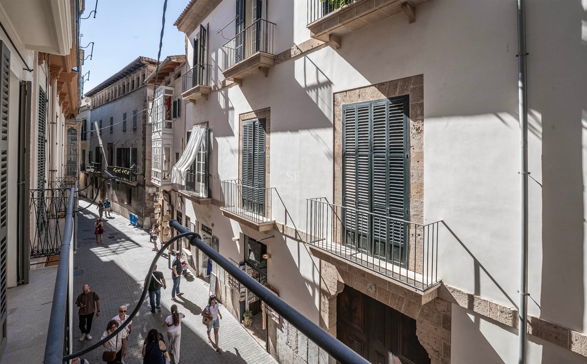 Vue depuis un balcon sur une rue étroite avec des bâtiments en pierre claire, des volets verts et des passants.