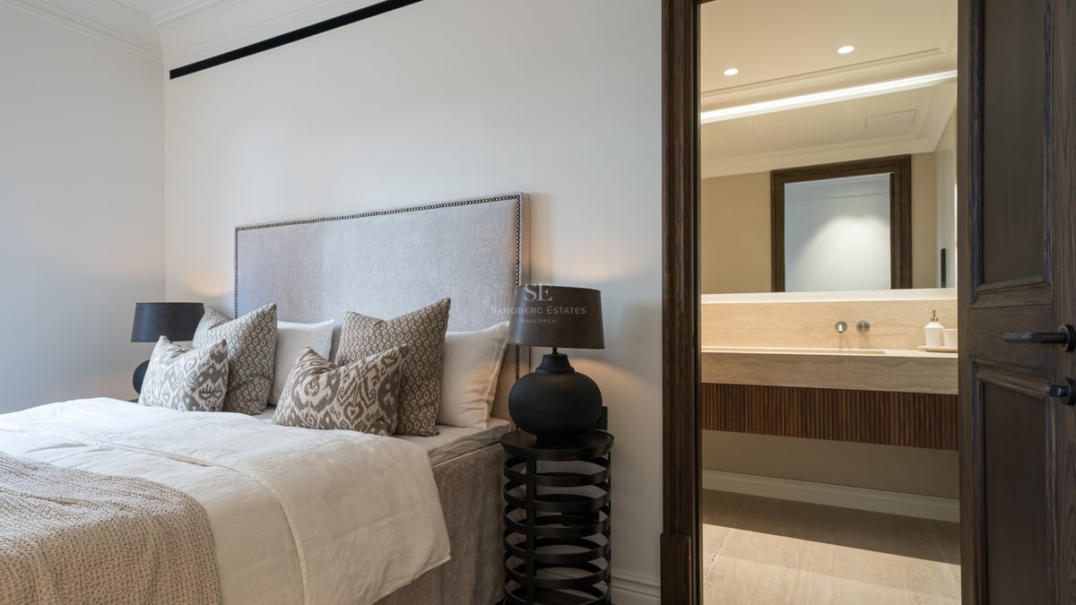 Modern bedroom featuring a tufted headboard and a view into a private bathroom with travertine stone vanity.