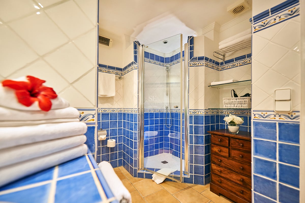 Salle de bain avec carrelage bleu et blanc, douche vitrée, sol en terre cuite et commode en bois.