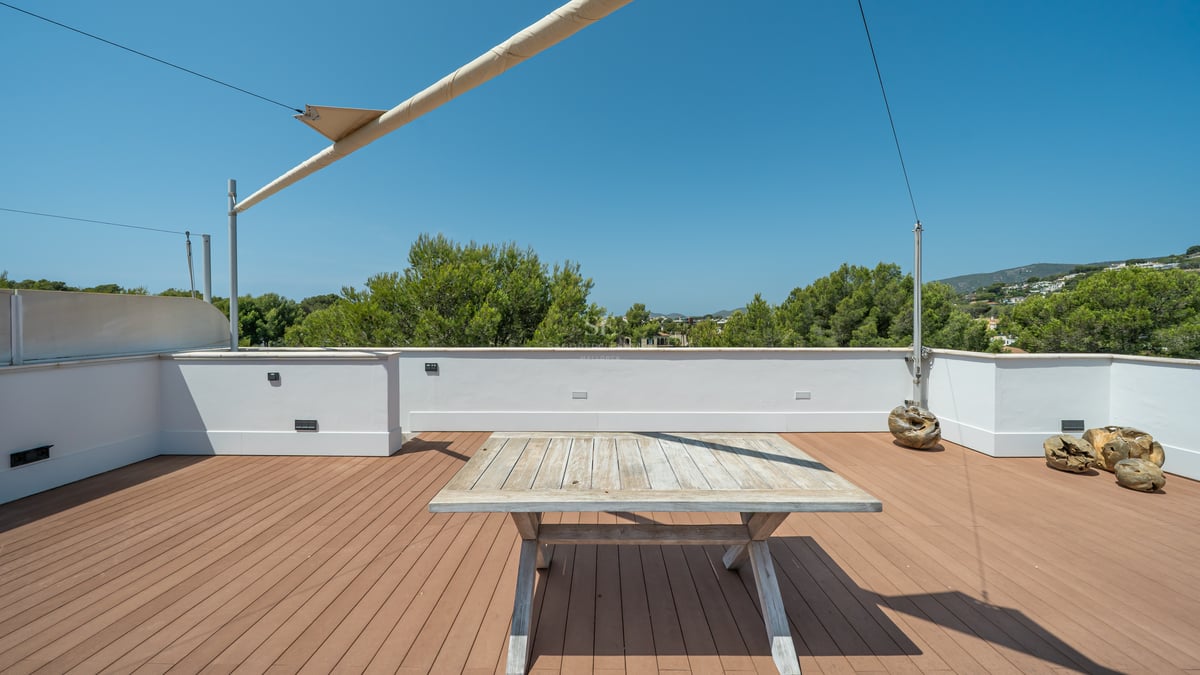 Grande terrasse de toit avec plancher en bois, table rustique, voile d'ombrage et vue sur les pins et collines.