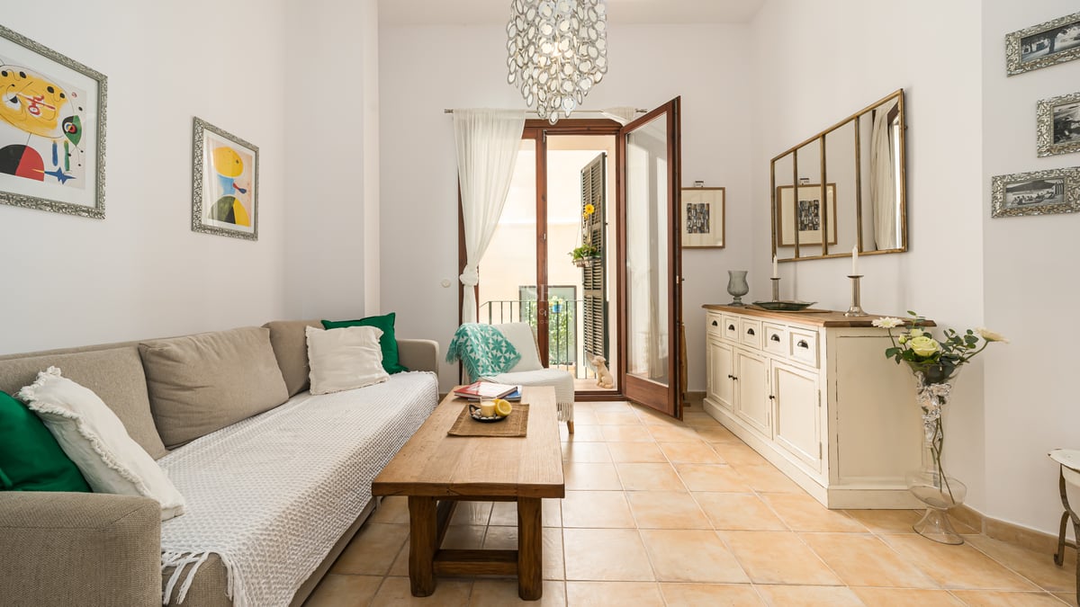 Salon baigné de lumière avec sol en terre cuite, canapé beige, table en bois et portes-fenêtres ouvrant sur un balcon.