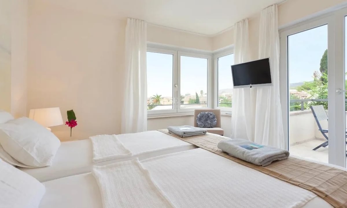 Chambre moderne avec deux lits, draps blancs, grandes fenêtres et porte vitrée ouvrant sur une terrasse ensoleillée.
