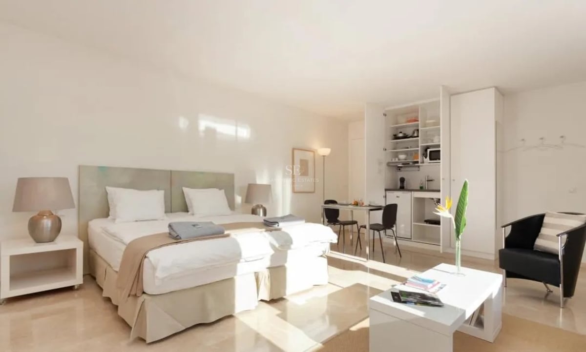 Studio lumineux avec lit blanc, sol en marbre et kitchenette dissimulée dans des placards blancs.