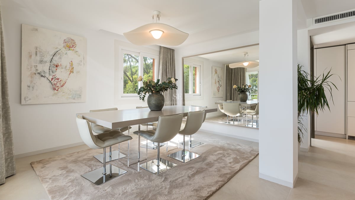 Salle à manger moderne avec table aux tons neutres, chaises chromées, grand miroir mural et vue sur le jardin.