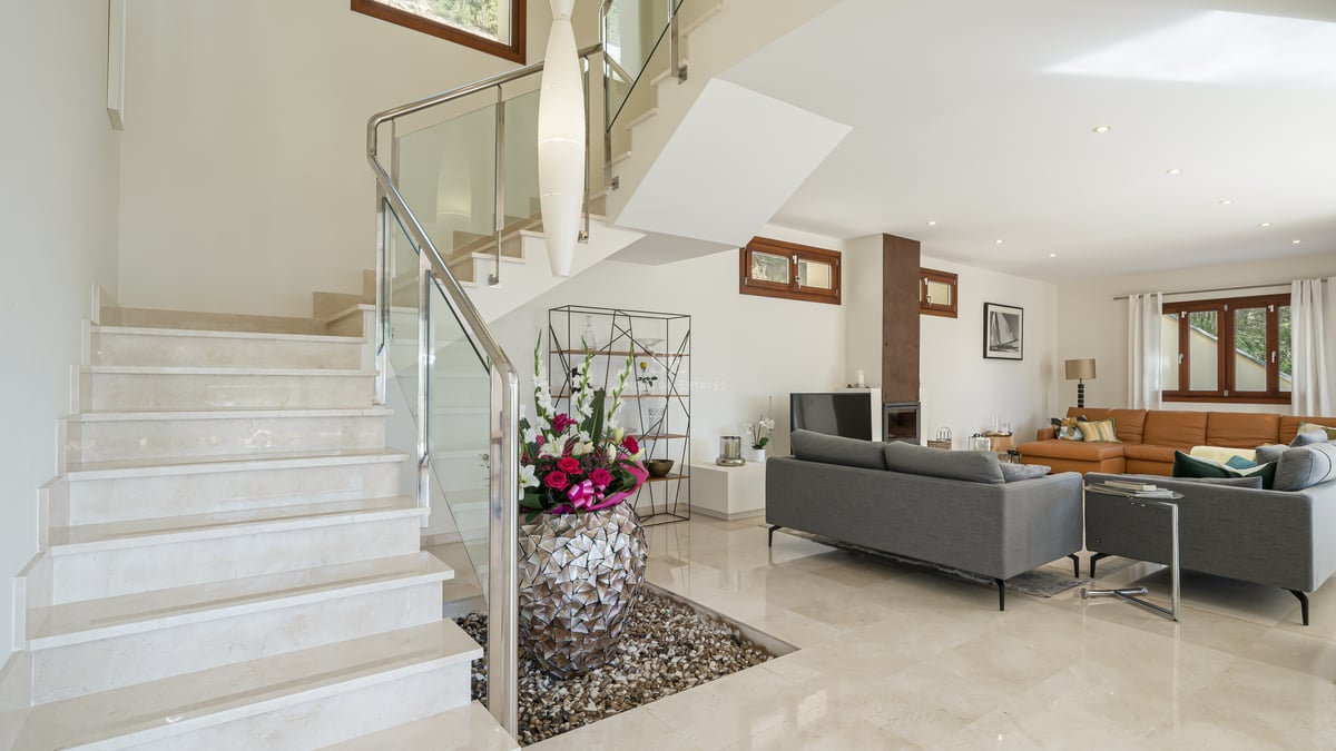 Salon lumineux à aire ouverte avec escalier en marbre crème, balustrades en verre et canapés gris.
