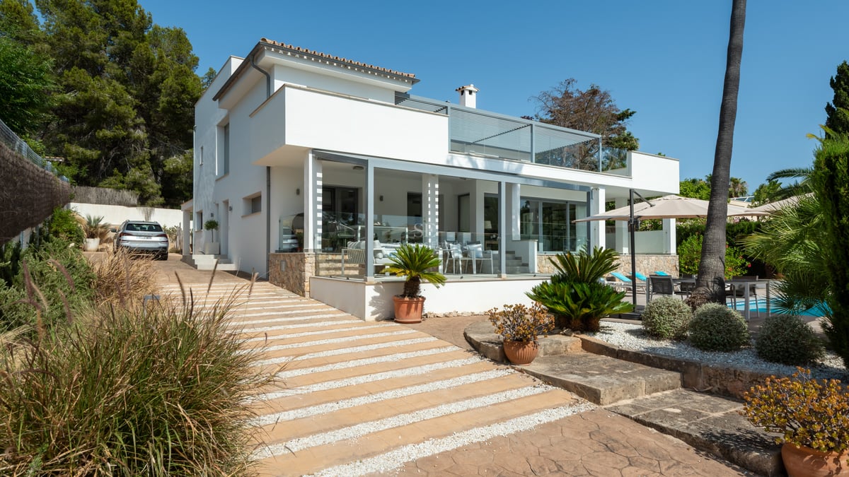 Villa blanche moderne de deux étages avec terrasses vitrées, accents en pierre et jardin soigné sous un ciel bleu clair.