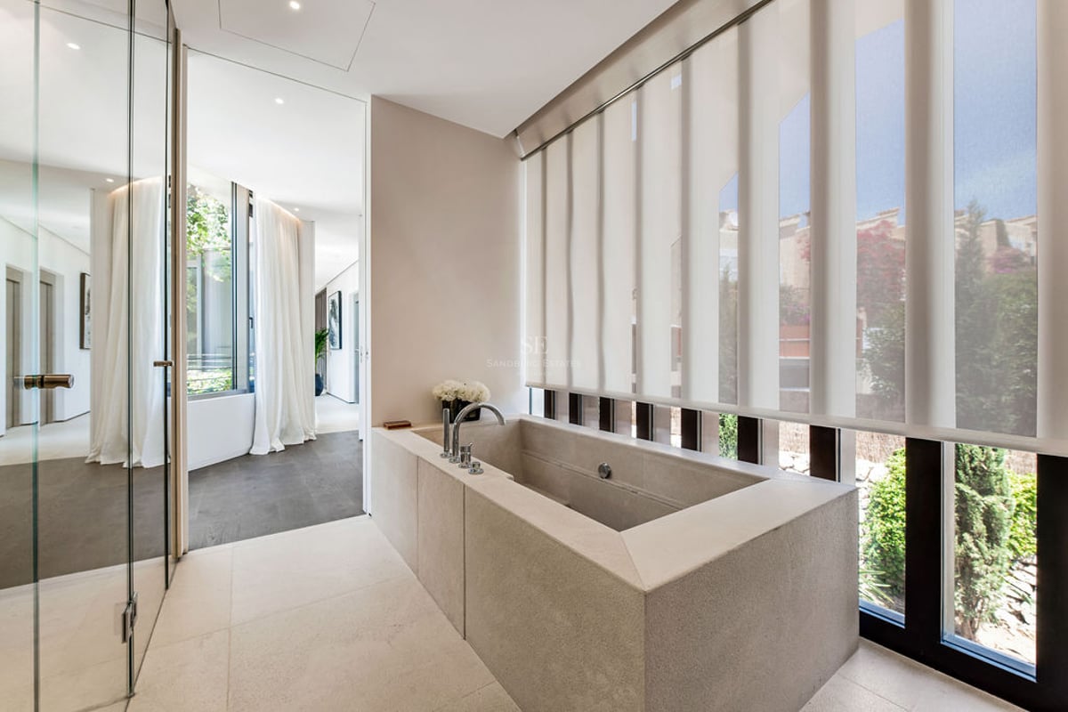 Salle de bain minimaliste avec baignoire rectangulaire en pierre, parois vitrées et grandes fenêtres.