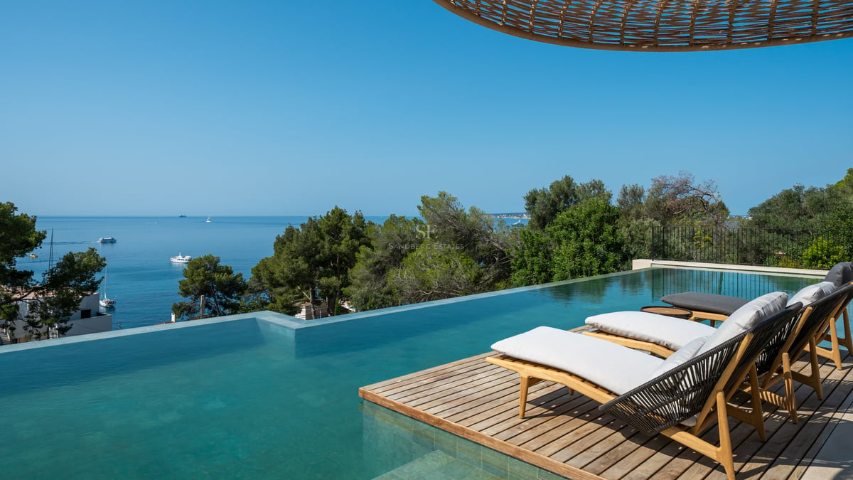 Piscine à débordement avec terrasse en bois et bains de soleil face à la mer bleue et aux pins.